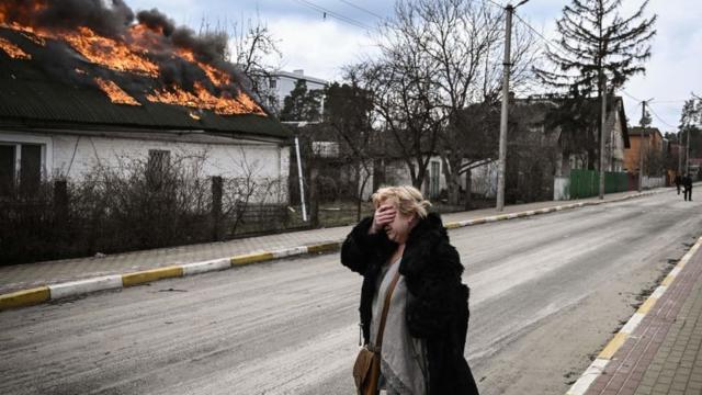 A woman covers her face with her hand as she stands in front of a house burning in Irpin, outside Kyiv, 4 March 2022