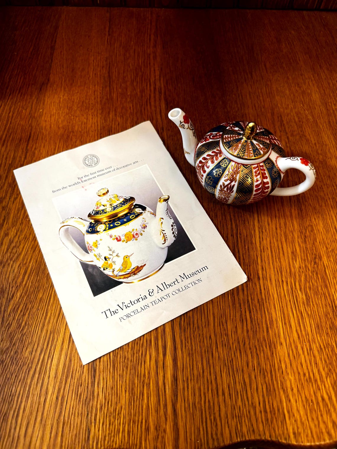 May include: A decorative porcelain teapot from the Victoria & Albert Museum Porcelain Teapot Collection. The teapot has a gold lid and handle, with a colorful pattern of red, blue, and gold. A brochure with a picture of a teapot is next to it.