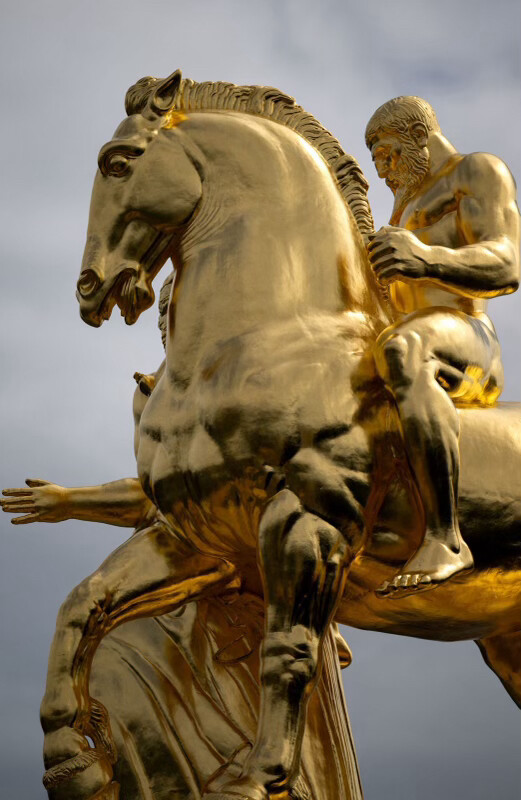 Golden statue of a muscular man riding a horse, with intricate details and a cloudy sky in the background.