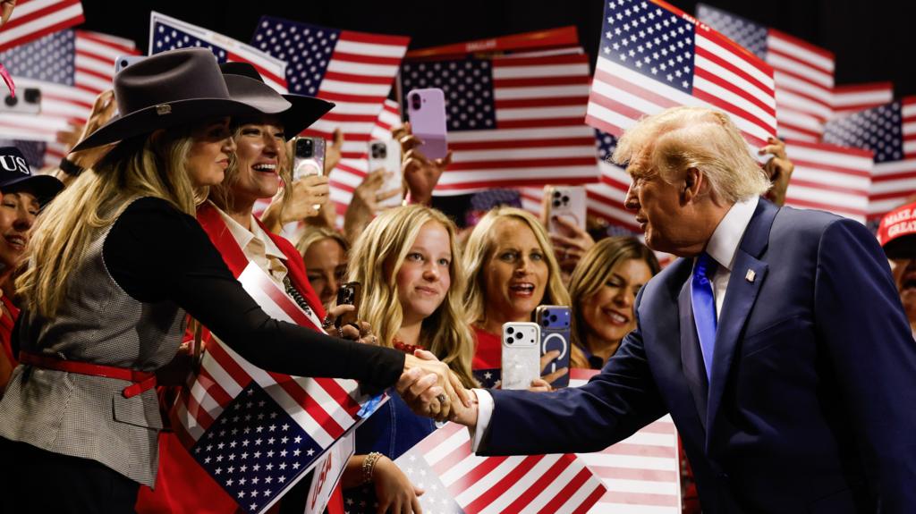 Donald Trump shakes hands with a woman wearing a cowboy hat in front of cheering supporters